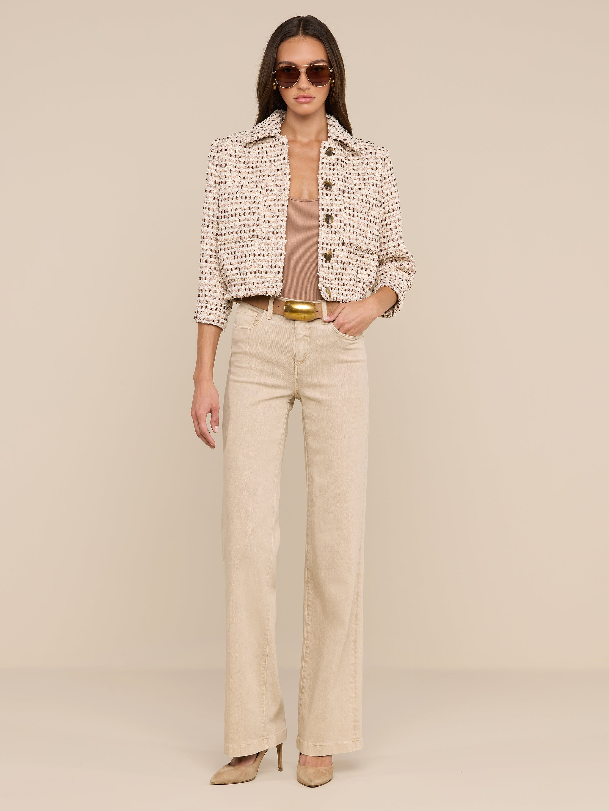A woman wearing sunglasses, the Jia Cropped Tweed Jacket with three-quarter sleeves, a tan top, wide-leg beige pants, and heels stands against a plain white background with her hands in her pockets.