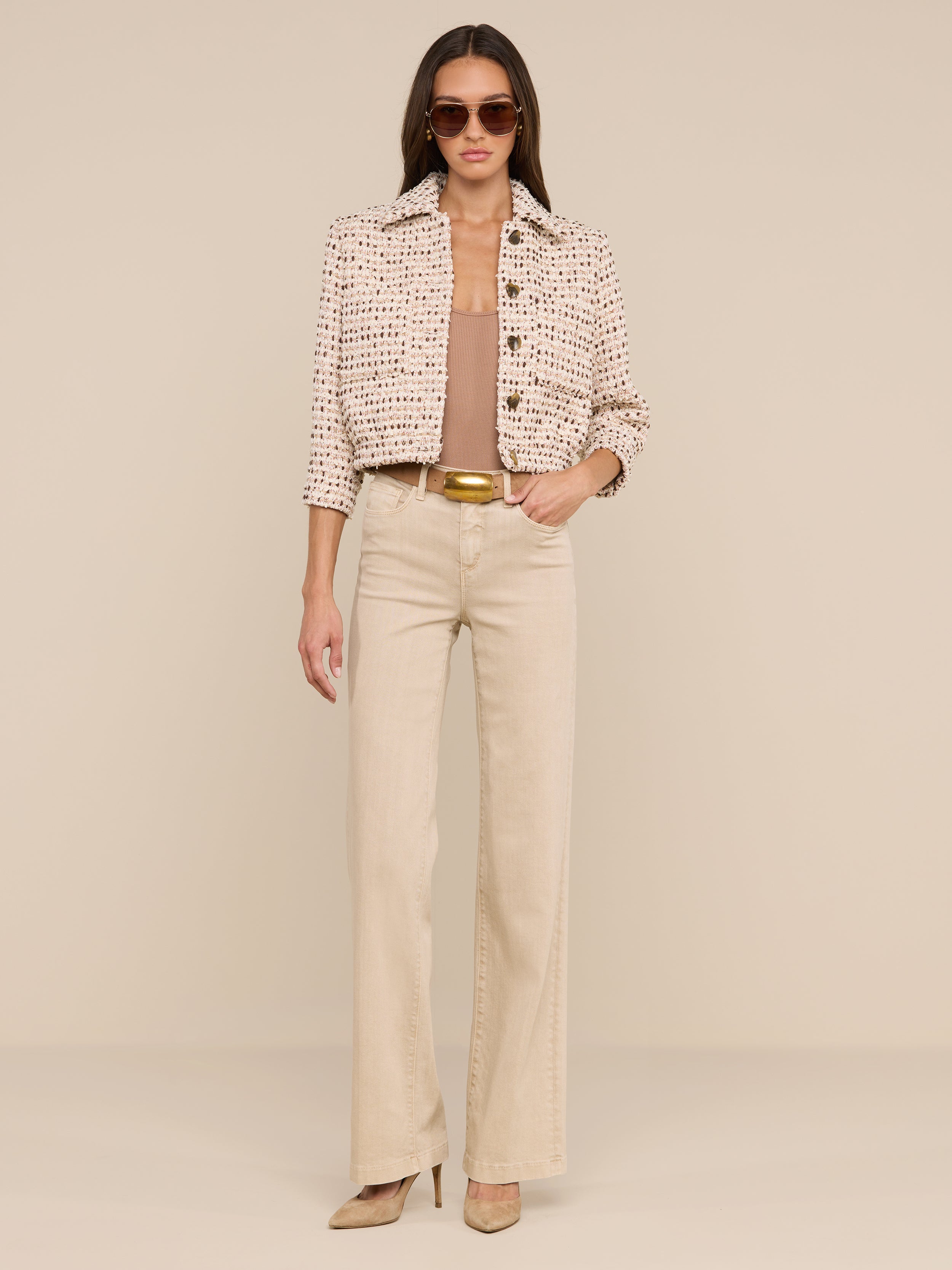 A woman wearing sunglasses, the Jia Cropped Tweed Jacket with three-quarter sleeves, a tan top, wide-leg beige pants, and heels stands against a plain white background with her hands in her pockets.