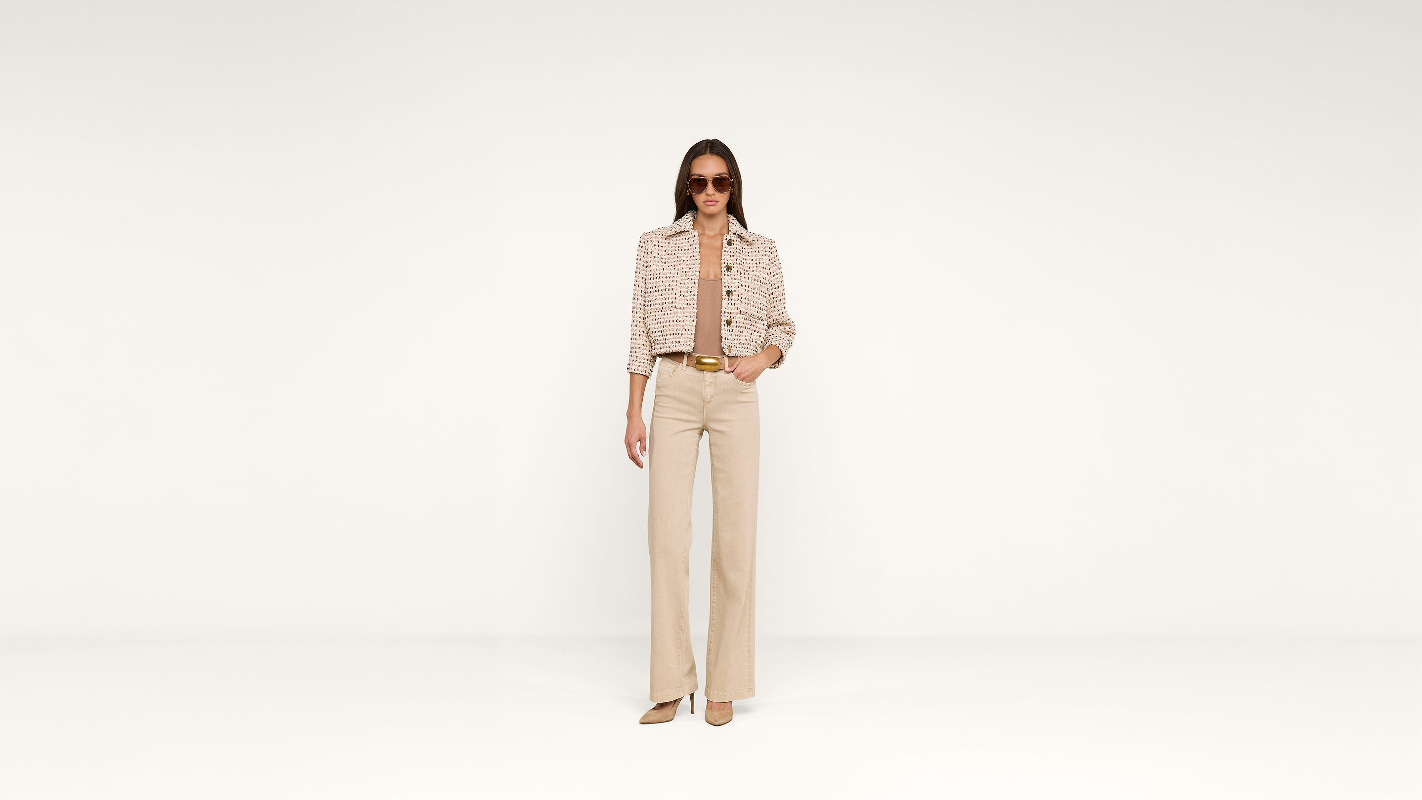 Woman stands against a plain white background wearing sunglasses, a patterned jacket, beige high-waisted pants, and matching heels, with her hands in her pockets.