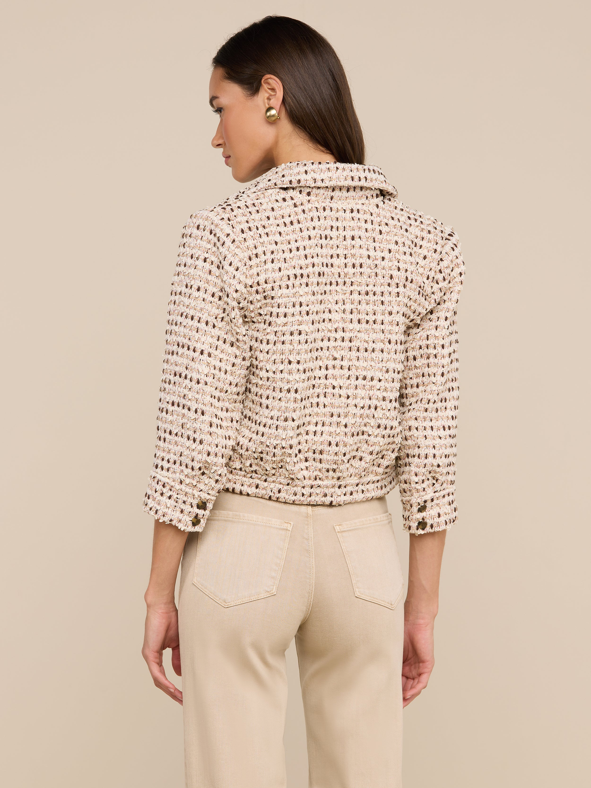 A woman with straight hair tucked behind her ear and round gold earrings stands with her back facing the camera. She is wearing an ivory Jia Cropped Tweed Jacket featuring three-quarter sleeves, paired with beige pants. The background is plain and light.