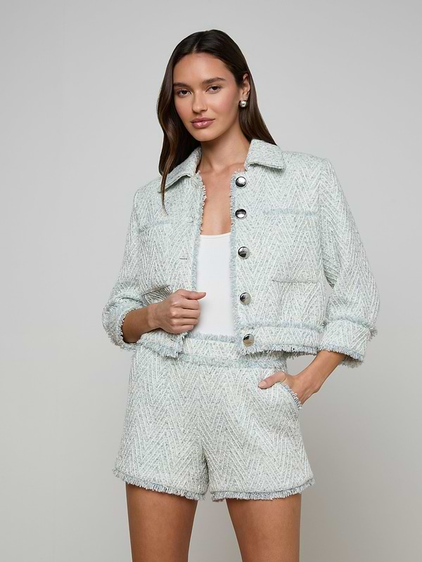 A woman wears the Jia Fringed Tweed Jacket in light blue chevron with matching shorts over a white top, standing with one hand in her pocket and the other holding the jacket, against a plain light-colored background.