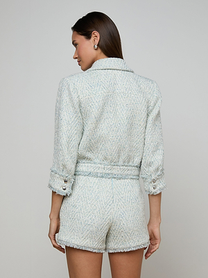 A woman faces away from the camera, wearing the Jia Fringed Tweed Jacket in light blue and white chevron with rolled-up sleeves, micro sequin accents, and charming fringed trim, paired with matching high-waisted shorts.
