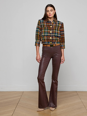 A woman stands indoors on a wooden floor against a white wall, wearing the Jia Cropped Tweed Jacket with gold buttons, styled with brown flared leather pants and beige pointed-toe shoes.
