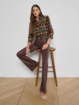 A woman with long brown hair sits on a wooden stool against a white wall, wearing the Jia Cropped Tweed Jacket, brown leather pants, and beige heels, with one leg bent and the other extended.