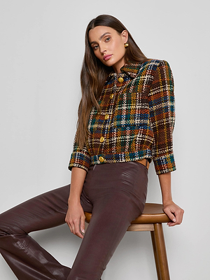 A woman with long brown hair sits on a wooden stool, wearing the Jia Cropped Tweed Jacket and high-waisted brown leather pants. She gazes at the camera with a relaxed expression.