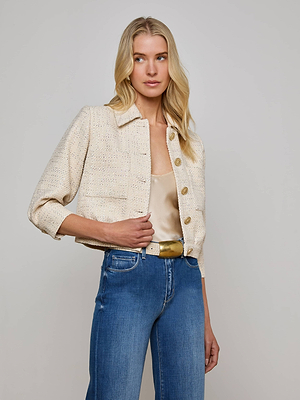 A woman with long blonde hair wears the Jia Sequined Tweed Jacket by L'AGENCE Sale over a beige satin top, paired with blue high-waisted jeans and a yellow belt, standing against a plain light gray background.