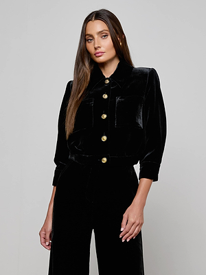 A woman with long, straight brown hair wears the L'AGENCE Jia Velvet Jacket with three-quarter sleeves and gold buttons, paired with matching black pants, standing against a plain light gray background.