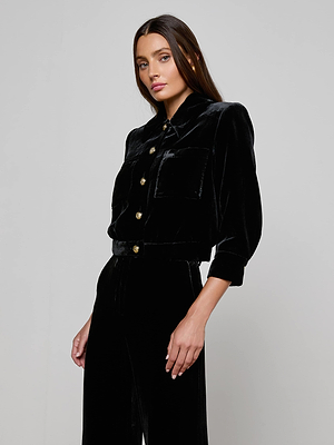 A woman with long brown hair models the L'AGENCE Jia Velvet Jacket—black, cropped, and featuring chest pockets—paired with matching high-waisted pants against a plain light gray background.