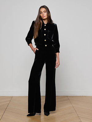 A woman with long brown hair stands on a wooden floor, wearing the L'AGENCE Jia Velvet Jacket—black, cropped, with large gold buttons—and matching wide-leg pants, set against a plain light background.