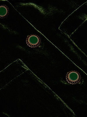 A close-up of the Jia Velvet Jacket by L'AGENCE in olive green, featuring gold and green decorative buttons and a partial pocket. The velvet texture and button details are clearly visible.