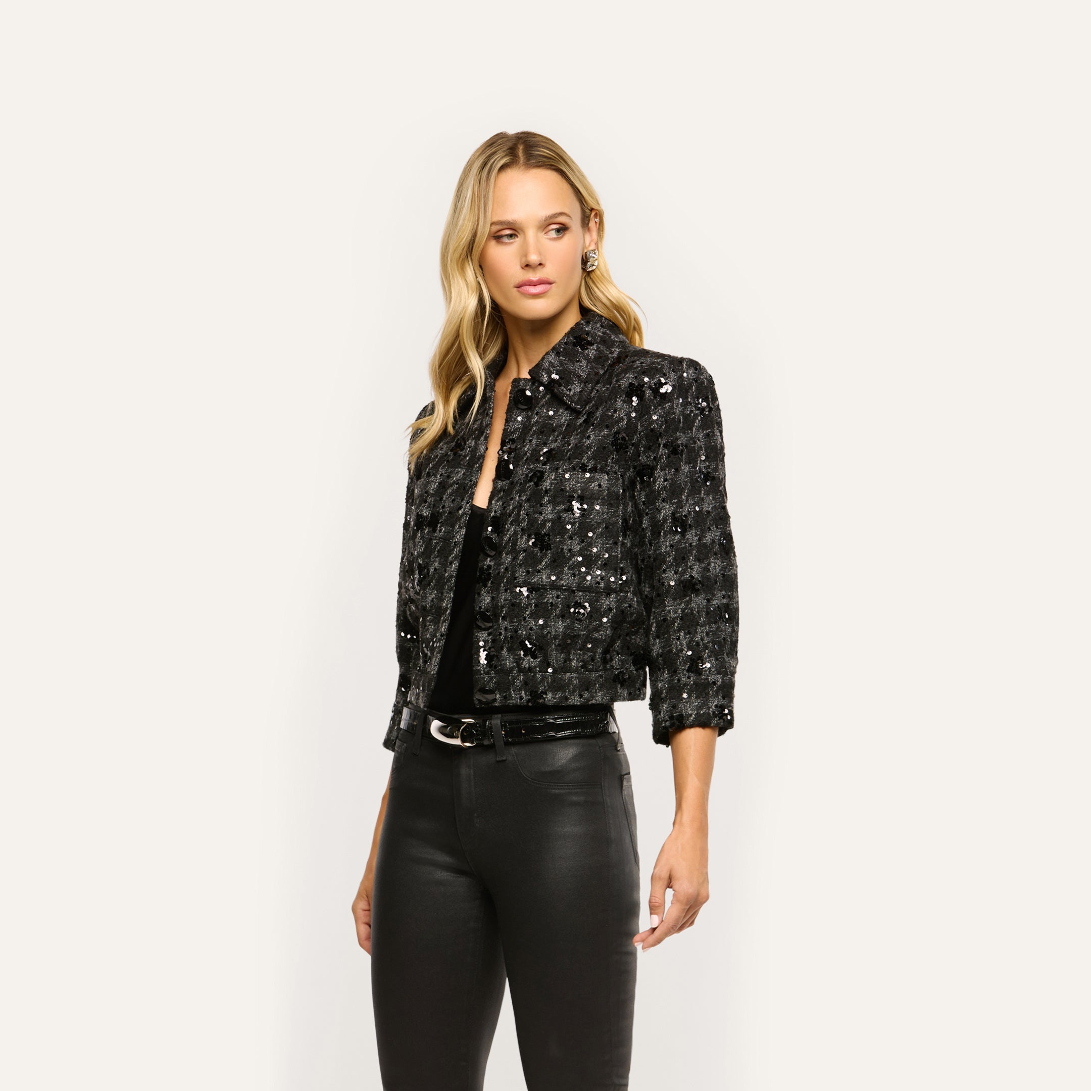 A woman wears a black, cropped Jia Sequined Tweed Jacket over a black top and pants.
