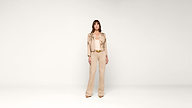 A woman stands facing forward, wearing a beige embroidered jacket, matching top, wide-legged pants, and tan heels, against a plain white background.