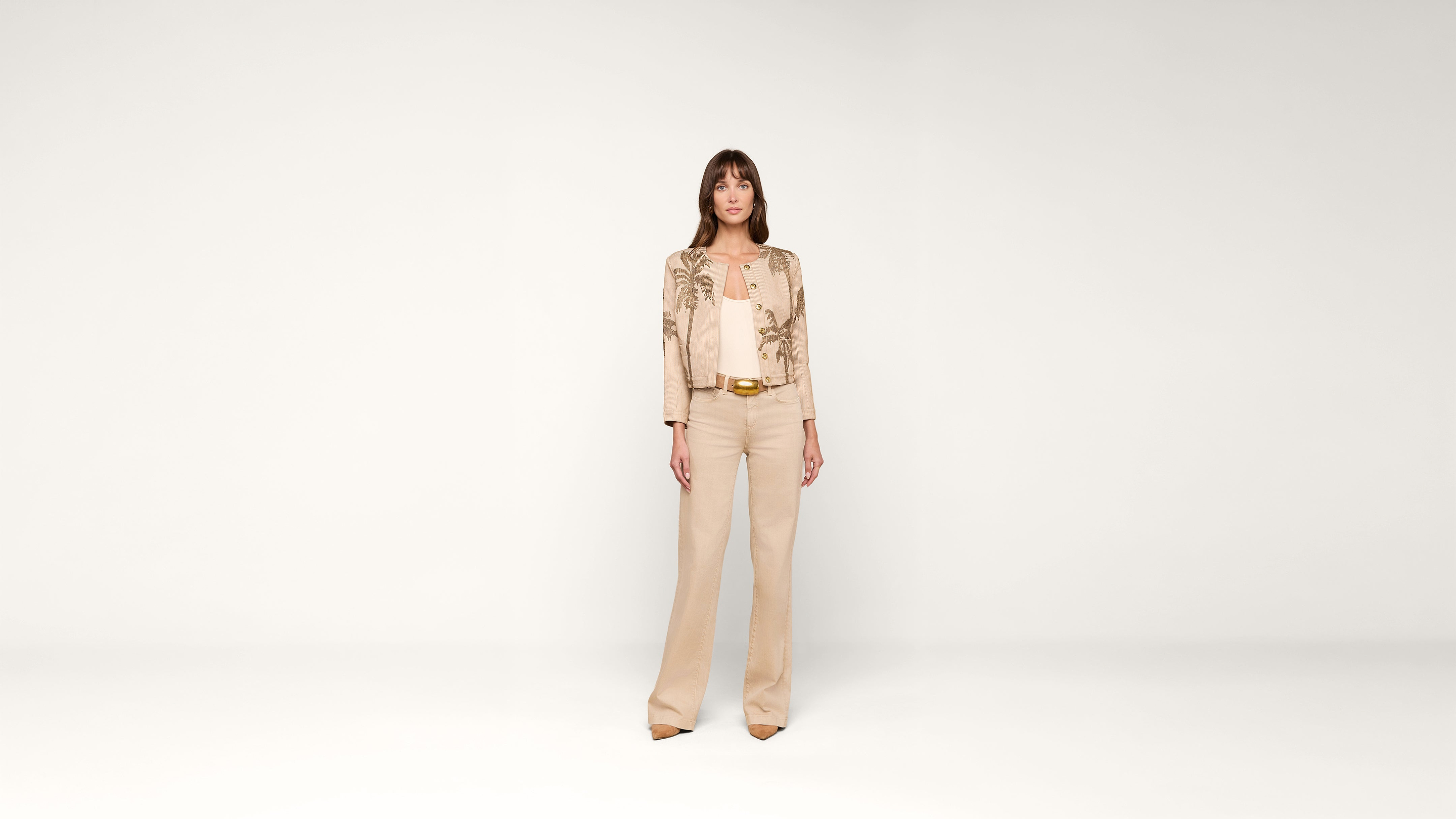 A woman stands facing forward, wearing a beige embroidered jacket, matching top, wide-legged pants, and tan heels, against a plain white background.