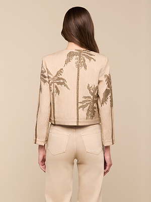 A woman with long brown hair stands with her back to the camera against a beige background, wearing the collarless Lian Beaded Jacket featuring palm tree motifs and matching beige pants.