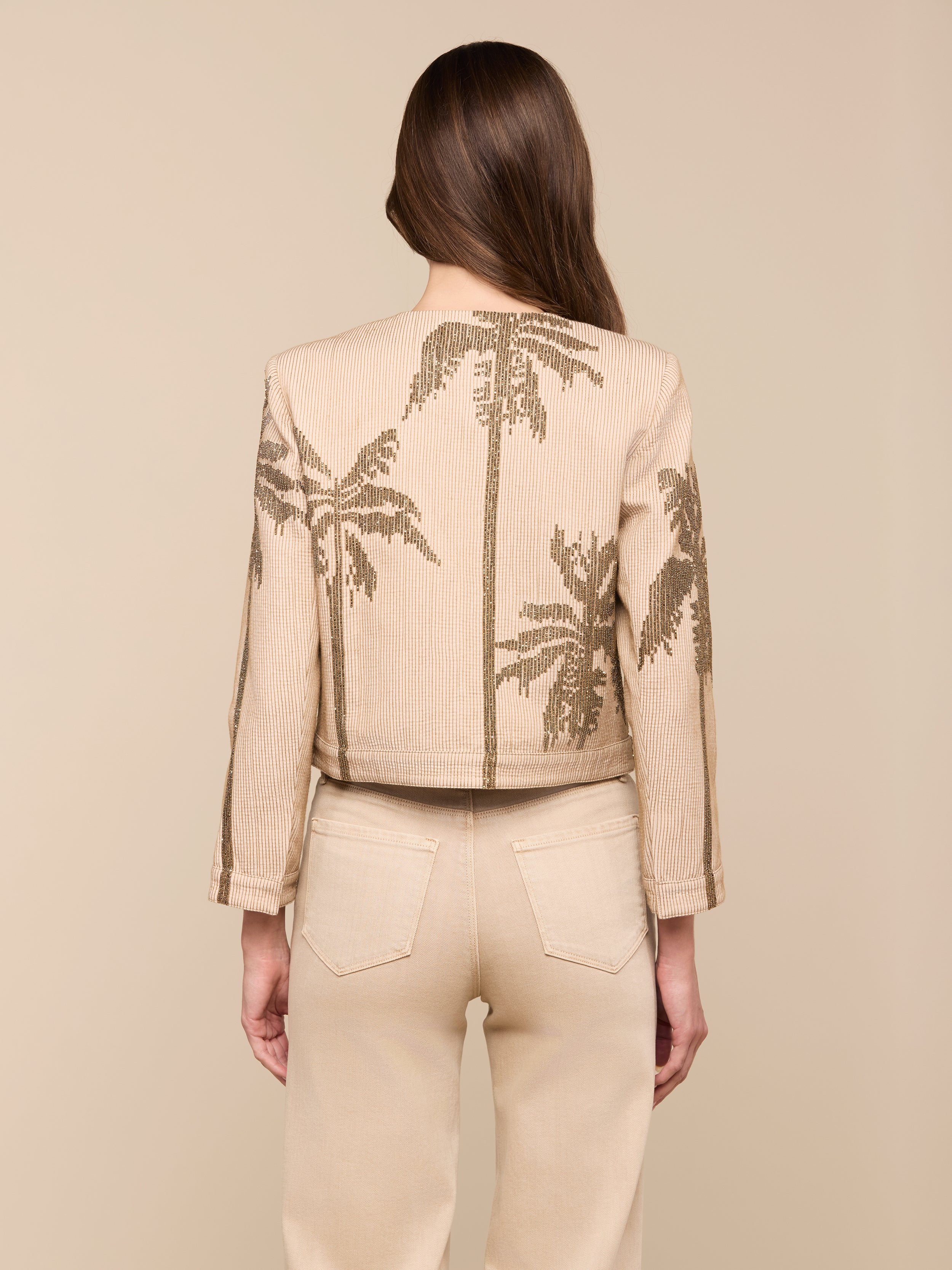 A woman with long brown hair, seen from behind, wears the Lian Beaded Jacket—a light beige, collarless piece featuring a palm tree motif—paired with matching high-waisted pants, set against a plain off-white background.
