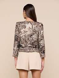 Woman facing away, wearing the Lian Tapestry Jacket crafted in Italian jacquard with scenic nature illustrations and light beige shorts, standing against a plain off-white background.