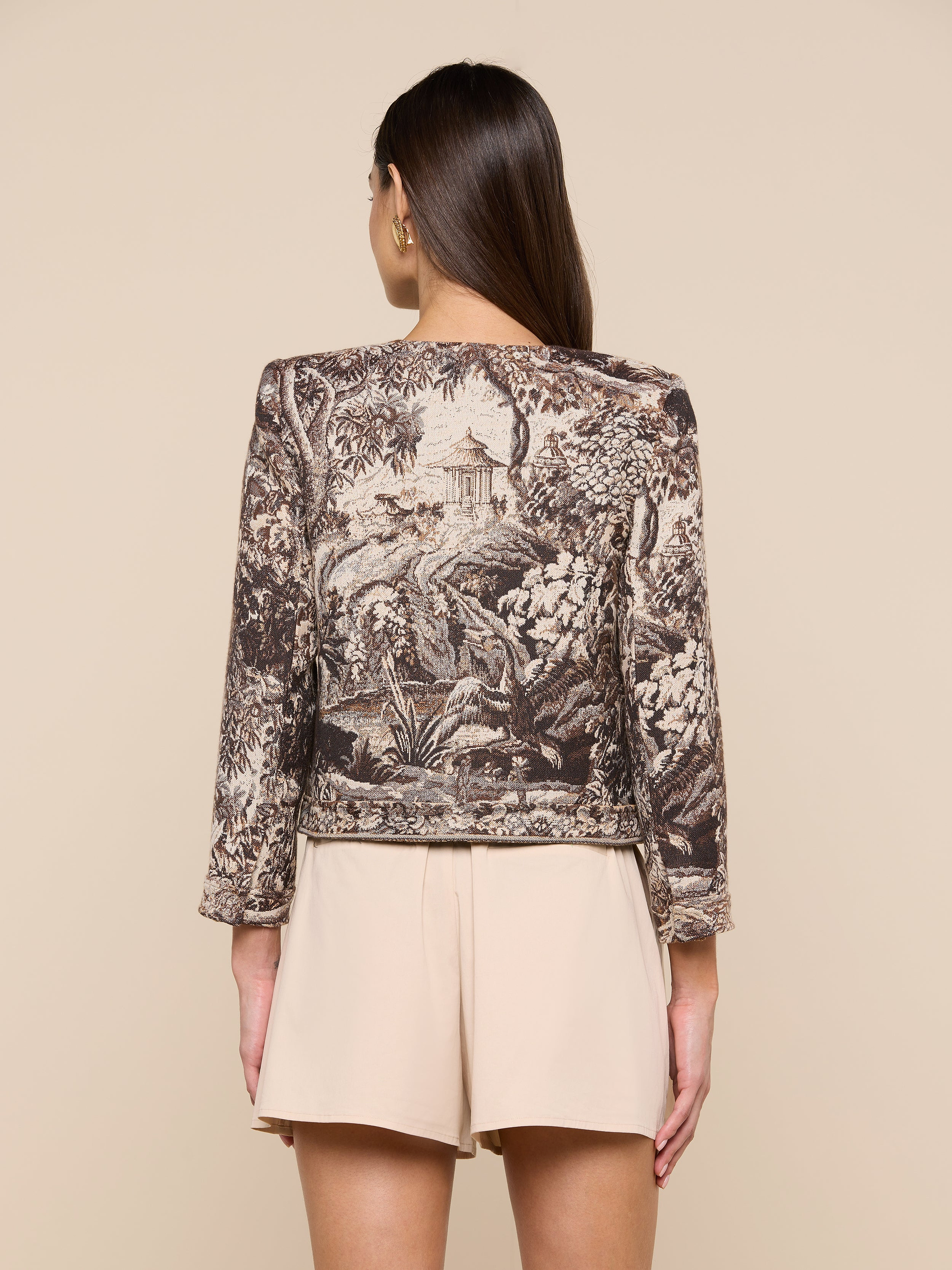 Woman facing away, wearing the Lian Tapestry Jacket crafted in Italian jacquard with scenic nature illustrations and light beige shorts, standing against a plain off-white background.