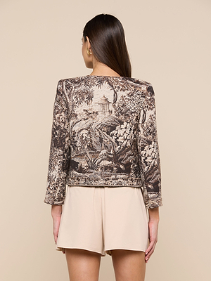 A woman with long brown hair, seen from behind, wears the Lian Tapestry Jacket—crafted from Italian jacquard with a scenic landscape—paired with high-waisted light beige shorts against a plain beige background.
