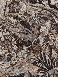 Close-up of the intricately woven fabric from the Lian Tapestry Jacket, featuring a mix of brown, black, and white threads that create a complex, abstract, and textured pattern. Folded sections introduce diagonal lines and added depth to the design.