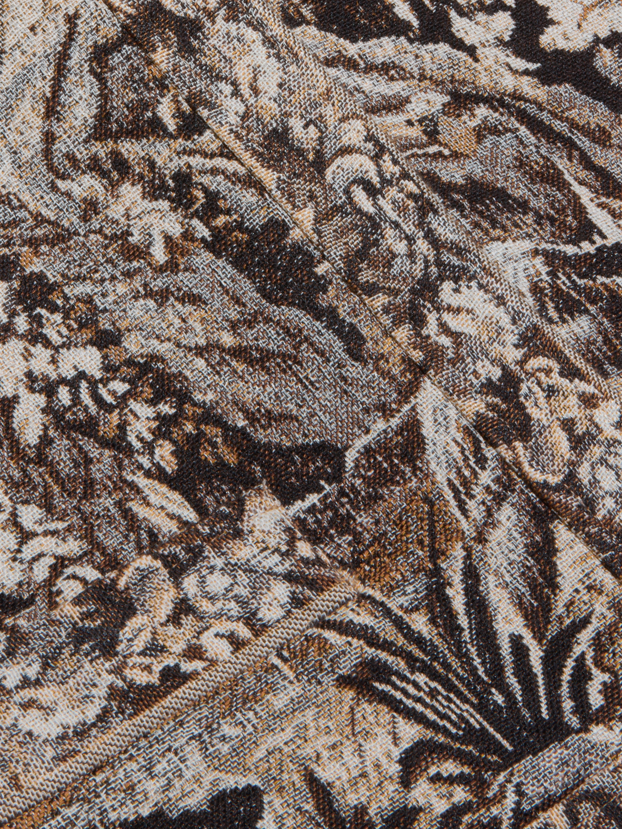 Close-up of the intricately woven fabric from the Lian Tapestry Jacket, featuring a mix of brown, black, and white threads that create a complex, abstract, and textured pattern. Folded sections introduce diagonal lines and added depth to the design.