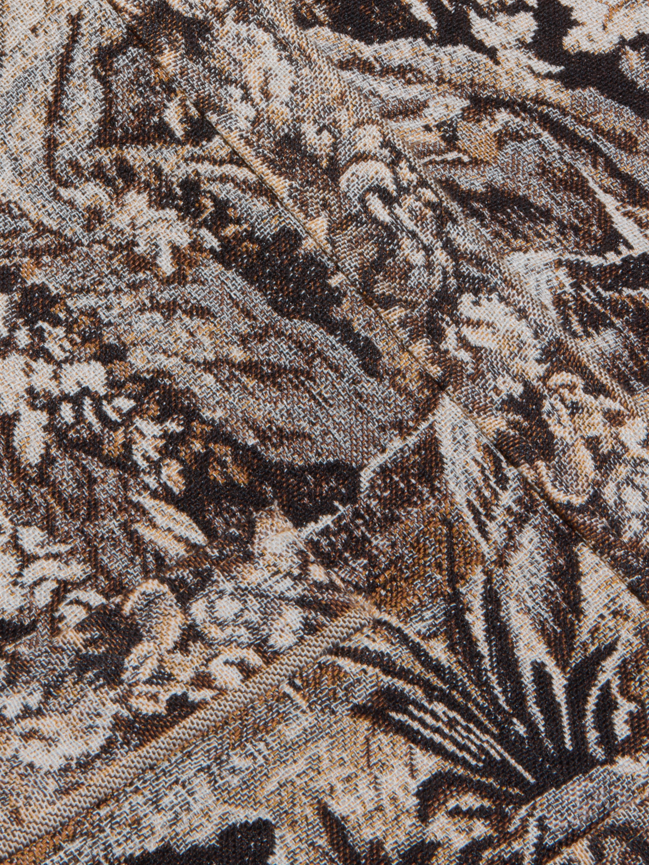 Close-up of the intricately woven fabric from the Lian Tapestry Jacket, featuring a mix of brown, black, and white threads that create a complex, abstract, and textured pattern. Folded sections introduce diagonal lines and added depth to the design.