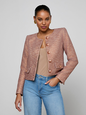 A woman poses confidently in the Zell Tweed Jacket, a collarless rose gold cropped style with gold buttons, paired with a beige top and light blue jeans against a plain light gray background.