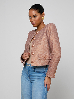 A woman wears the Zell Tweed Jacket, a collarless slub tweed piece in textured pinkish-beige, over a beige top and blue jeans. She pairs it with gold hoop earrings and has her hair pulled back, standing against a light gray background.