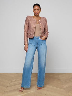 A woman stands against a plain wall wearing the Zell Tweed Jacket, a tan top, light blue high-waisted wide-leg jeans, and open-toe heels. She has one hand in her pocket and exudes confidence.