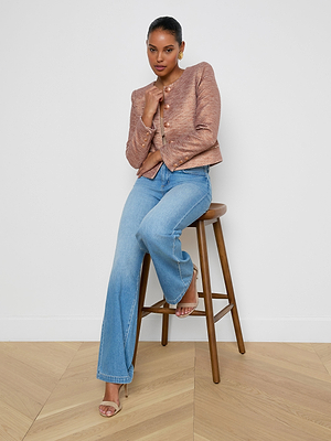 A woman wearing the Zell Tweed Jacket in textured copper sits on a wooden stool against a plain white wall, pairing it with light blue flared jeans, her legs crossed and one hand resting on her chin.