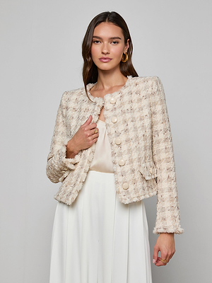 A woman with long brown hair wears the Zell Houndstooth Tweed Jacket over a light blouse and white skirt, standing against a plain light gray background and highlighting a sophisticated, feminine silhouette.