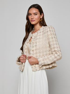 A woman with long brown hair wears the Zell Houndstooth Tweed Jacket over a white dress, standing against a plain light gray background, facing slightly to the side with a gentle expression.