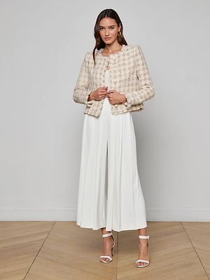 A woman poses on a wooden floor in the Zell Houndstooth Tweed Jacket, styled with a white pleated jumpsuit and white high-heeled sandals, highlighting a feminine silhouette against a plain light-colored wall.