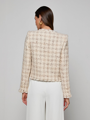 A woman with long brown hair, wearing gold hoop earrings and white pants, faces away from the camera against a light gray background, showcasing the Zell Houndstooth Tweed Jacket with a feminine silhouette.