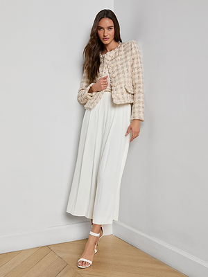 A woman with long brown hair wears the Zell Houndstooth Tweed Jacket over a flowing white dress and white heels, standing against a white wall on a wooden floor, looking at the camera, showcasing her feminine silhouette.