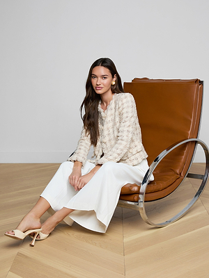 A woman with long brown hair sits on a modern brown leather chair, her silhouette highlighted by the Zell Houndstooth Tweed Jacket, white wide-leg pants, and beige heels against light wood floors and a plain white wall.