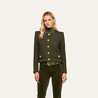A woman with long blonde hair wears the Zell Tweed Jacket—a collarless dark green piece with gold buttons—paired with green pants and a belt, standing against a plain light background.