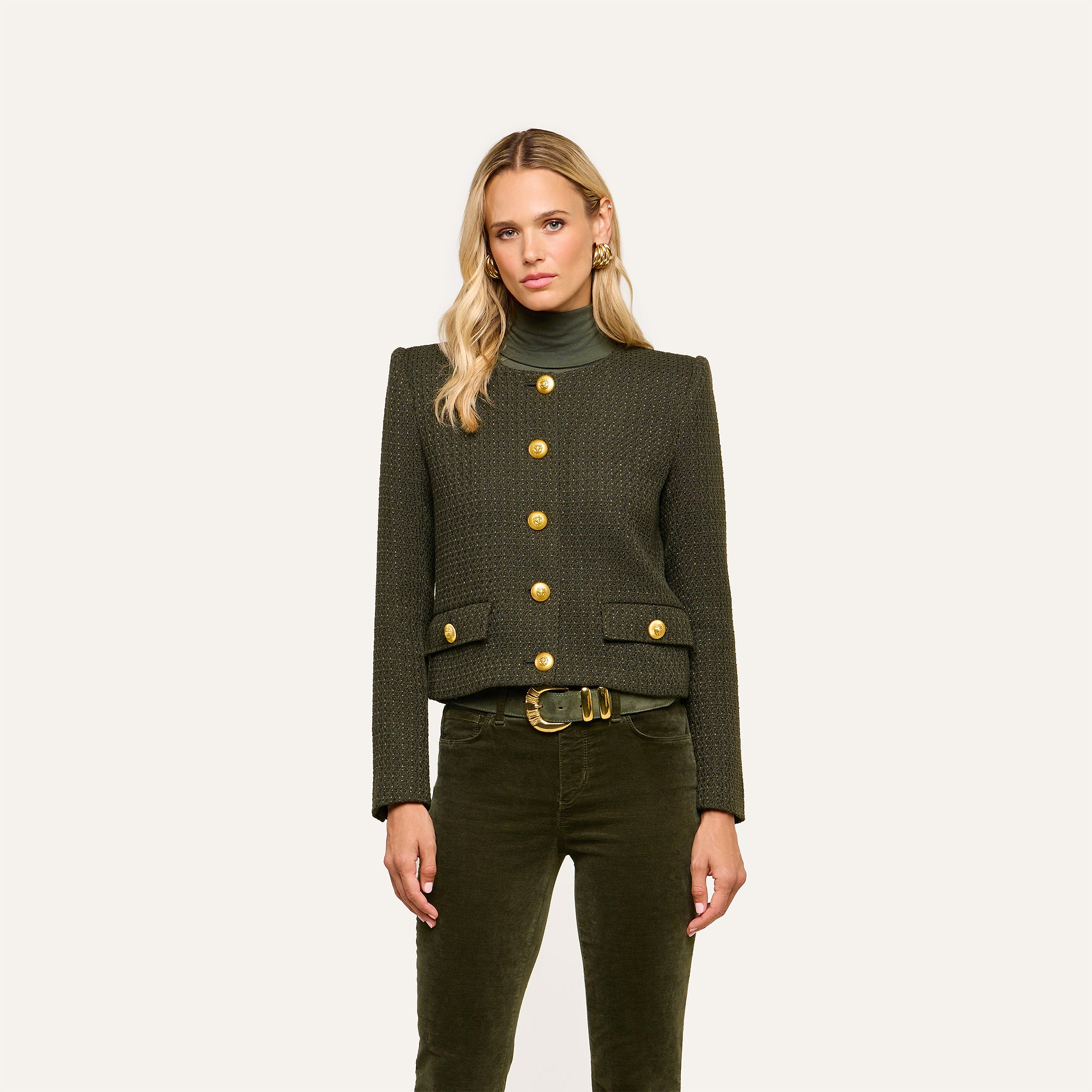 A woman with long blonde hair wears the Zell Tweed Jacket—a collarless dark green piece with gold buttons—paired with green pants and a belt, standing against a plain light background.