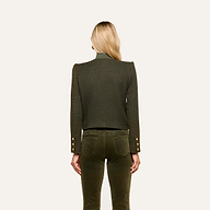 A woman with long blonde hair stands facing away, wearing the Zell Tweed Jacket—a textured, dark green tweed piece featuring gold buttons on the sleeves—paired with matching green pants. The background is plain and light-colored.
