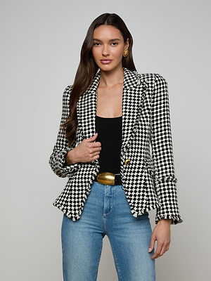 A woman with long brown hair wears the Kaisley Tweed Blazer in black and ivory over a black top, blue jeans, a wide gold belt, and gold hoop earrings, standing against a plain light background.