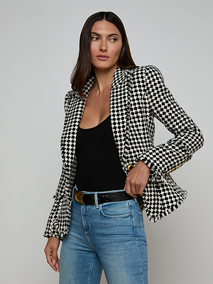 A woman stands against a plain background wearing light blue jeans, a black top, and the Kaisley Tweed Blazer in black and ivory, looking slightly to the side as she adjusts her blazer.