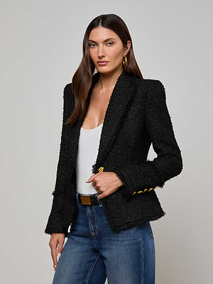 A woman with straight brown hair wears the L'AGENCE Kaisley Frayed Trim Blazer over a white top and blue jeans, standing forward against a plain light background with one hand holding her blazer.