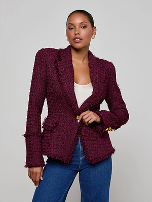 A woman poses confidently against a plain background wearing the L'AGENCE Kaisley Tweed Blazer over a white top and blue jeans, her hair pulled back with one hand on her waist.