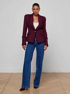 A woman stands against a plain light wall, wearing the L'AGENCE Kaisley Tweed Blazer over a white top, blue wide-leg jeans, and burgundy pointed shoes on a wooden floor.