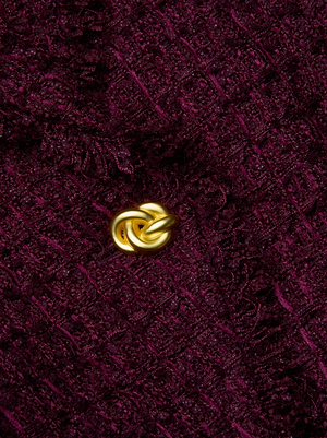A close-up of the textured, dark burgundy fabric from the L'AGENCE Kaisley Tweed Blazer, showcasing a single gold knot-shaped button at the center.