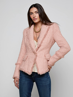 A woman with long brown hair models the L'AGENCE Kaisley Tweed Blazer in light pink over a cream top and blue jeans, standing with one hand on her hip against a simple light background.