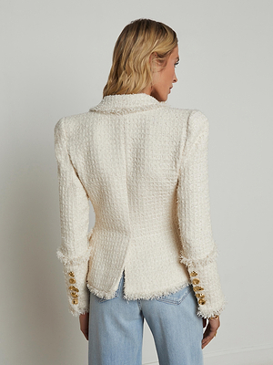A woman with blonde hair faces away, wearing the L'AGENCE Kaisley Tweed Blazer in cream with gold novelty buttons and light blue jeans, standing against a plain light-colored wall.