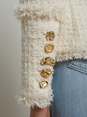 Close-up of a person wearing the L'AGENCE Kaisley Tweed Blazer in cream with frayed edges and four gold buttons on the sleeve, paired with light blue denim jeans.