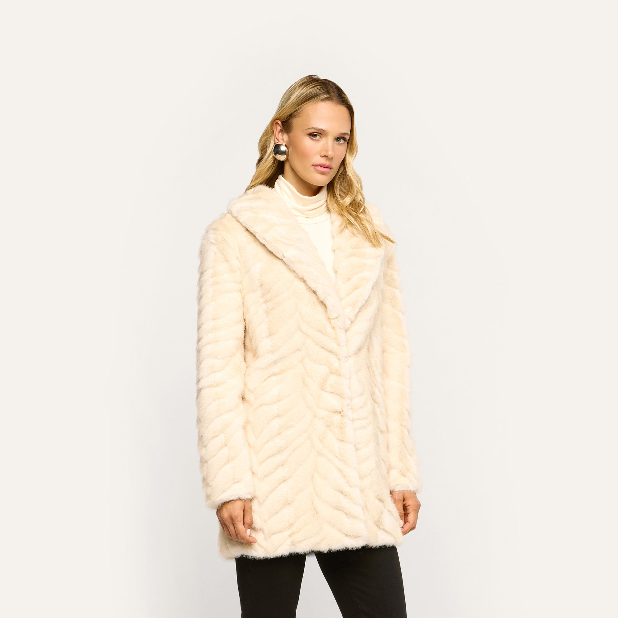 A woman wears a Toira Faux Fur Coat in a 70s-inspired cream chevron pattern over a turtleneck sweater.