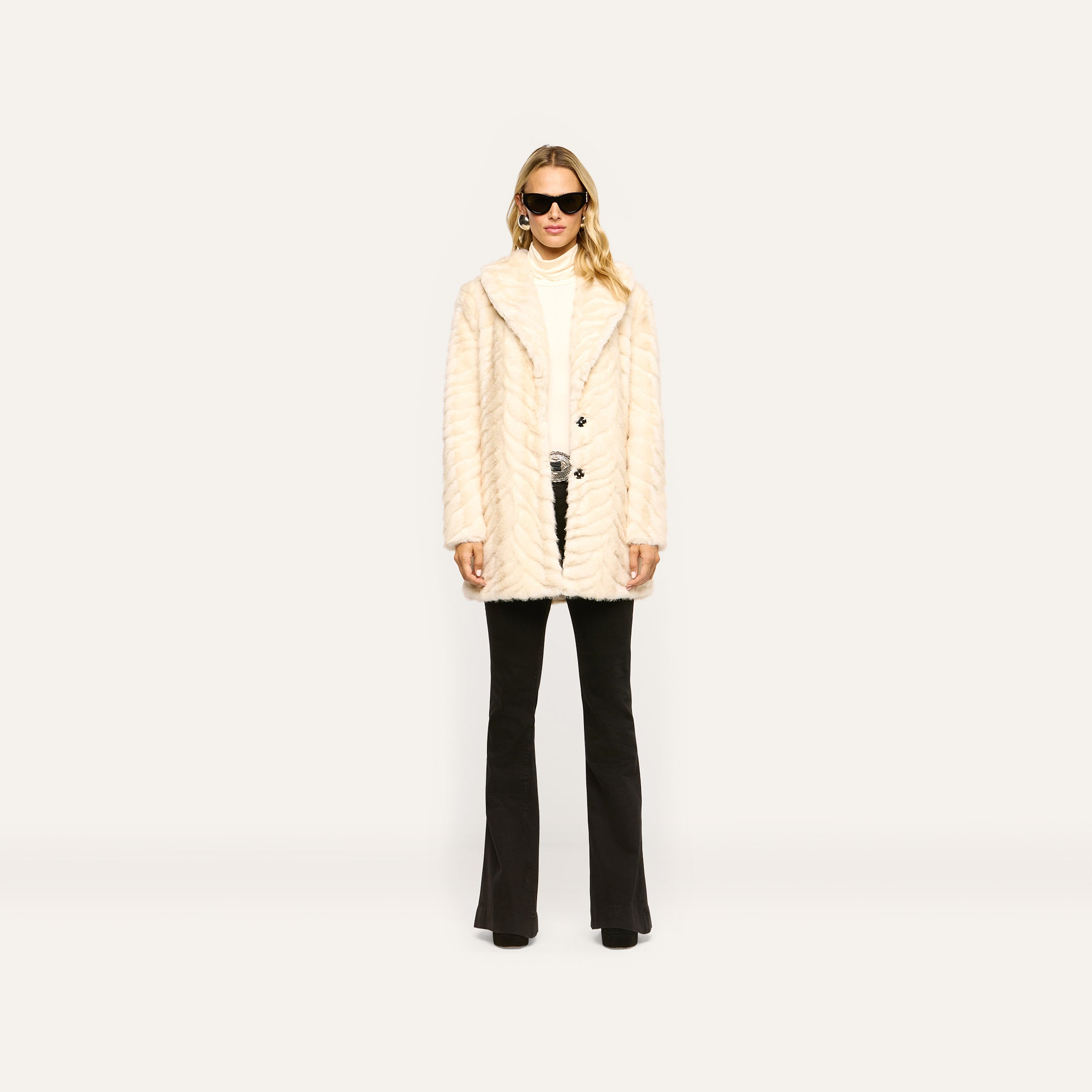 A person wearing a cream-colored Toira Faux Fur Coat in a 70s style over a white turtleneck and black flared trousers.