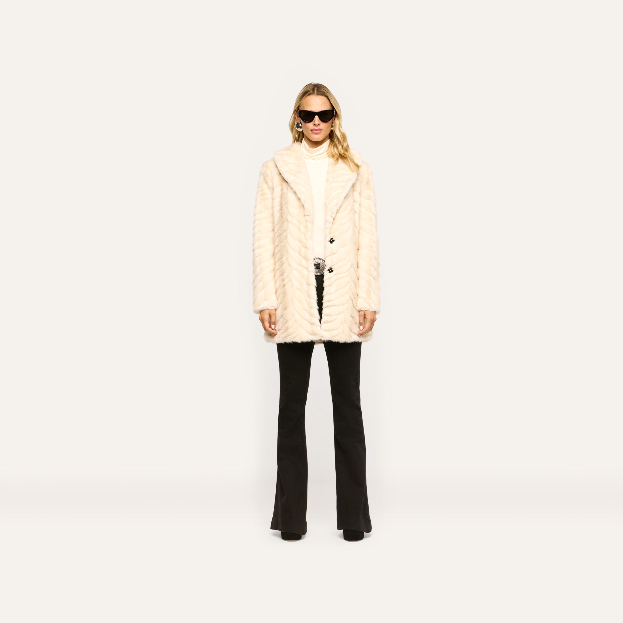 A person wearing a cream-colored Toira Faux Fur Coat in a 70s style over a white turtleneck and black flared trousers.